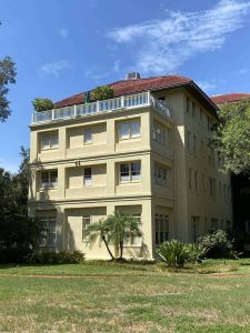 Spacious apartment building with a rooftop deck and lush landscaping, ideal for homebuyers seeking comfortable residential solutions. Modern multi-story residential building with a yellow exterior, large windows, and a rooftop terrace, surrounded by greenery under a clear blue sky.